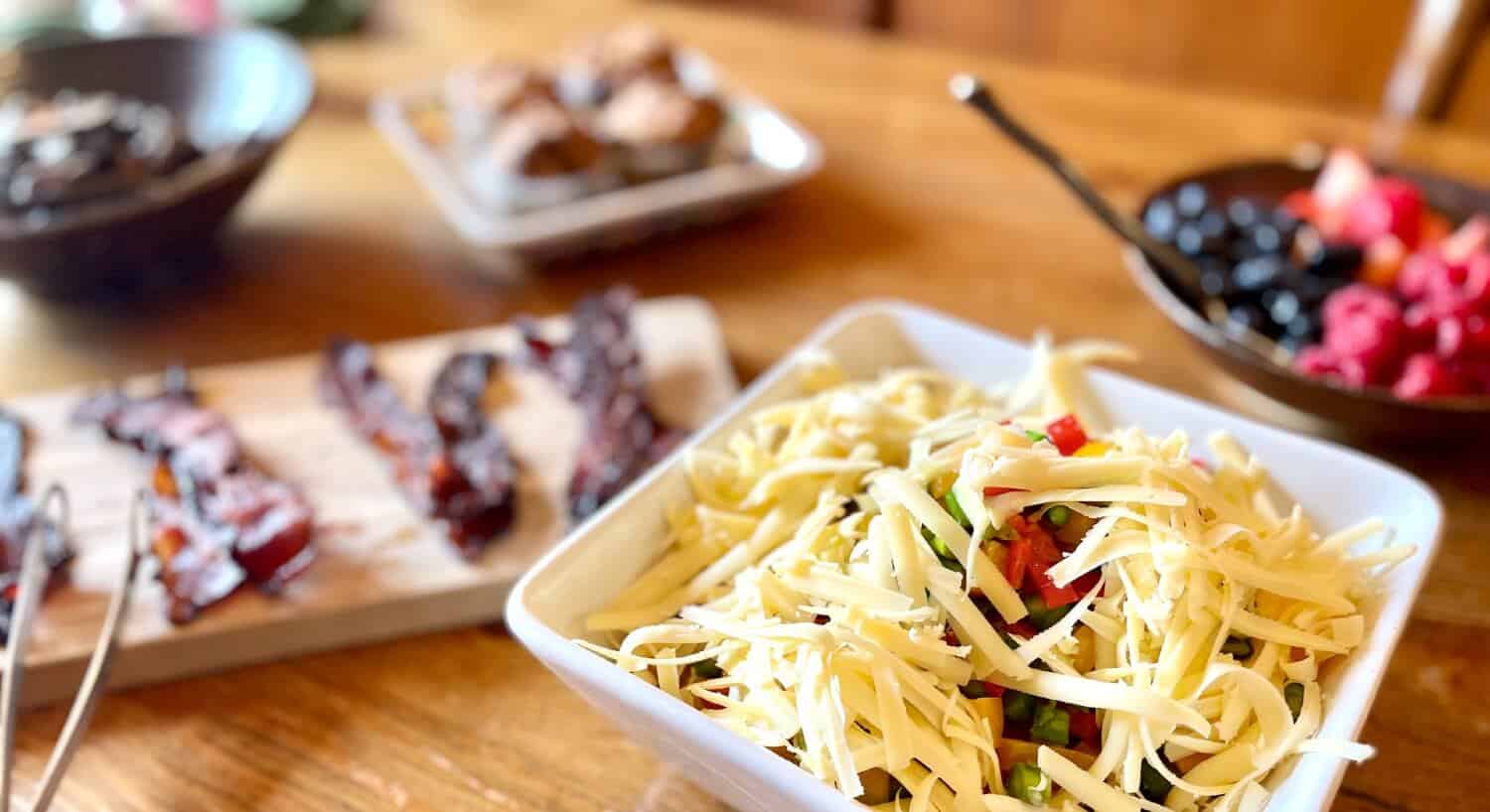 Breakfast table with bacon, bowl of berries, muffins and square white bowl with food topped with shredded cheese