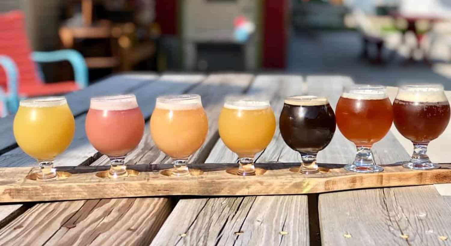 Wood picnic table with a flight of seven glasses of different colors of beer