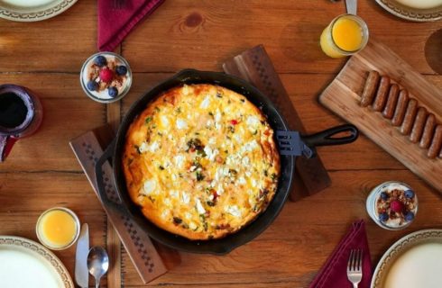 Wood dining able with cast iron pan with a breakfast dish, platter of sausage links and glasses of orange juice