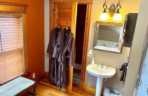 Bathroom with pedestal sink, robes hanging on a wood door, shower and bench underneath a window with blinds