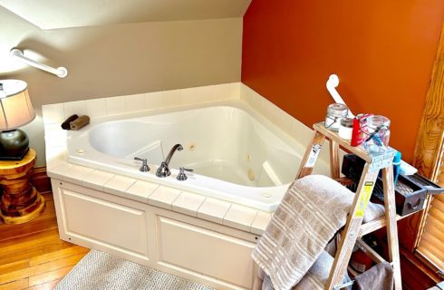 Corner jacuzzi tub next to a window with blinds and ladder holding towels and bath amenities
