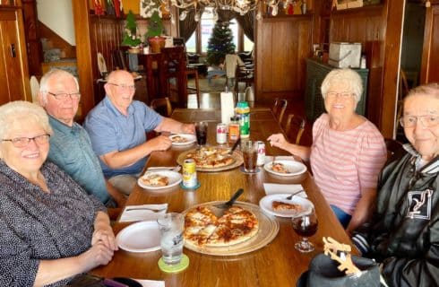 A group of five elderly people sitting at a dining table in a home eating pizza