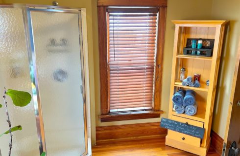 Bathroom with corner stand up shower with glass doors, window with blinds and shelving unit with towels