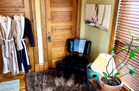 Bathroom with plush robes hanging on a door, leather chair, window with blinds and table with lamp and potted plant