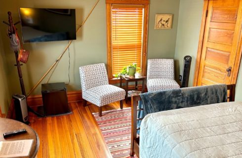 Bedroom with queen bed, corner TV and two sitting chairs and table by a window with blinds