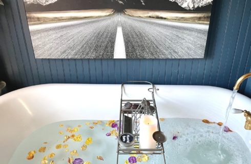 White clawfoot tub filled with water with flower petals and gold faucet underneath a painting