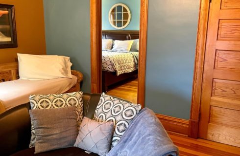 Sitting room of a bedroom with a bench, round club chair with pillows and doorway showing a bed