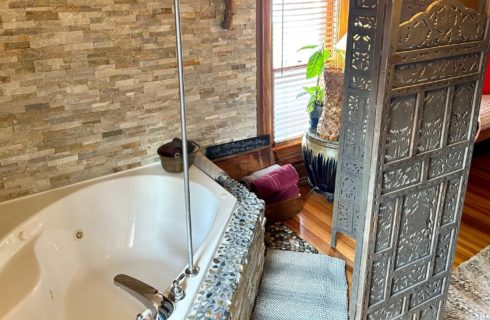 Corner jacuzzi tub with stone feature, privacy screen and potted plant by a window with blinds