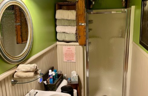 Small bathroom with stand up shower, pedestal sink, round silver-framed mirror and cubby with green and white towels