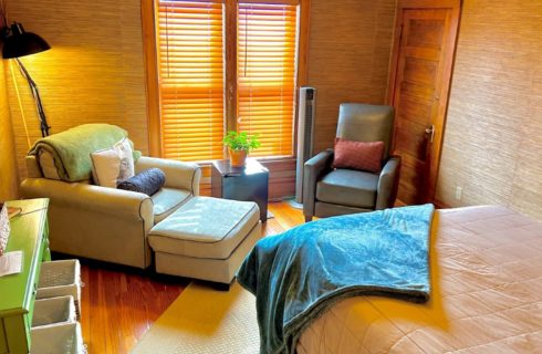 Bedroom with queen bed, natural grass wallpaper and two sitting chairs by a large window with blinds