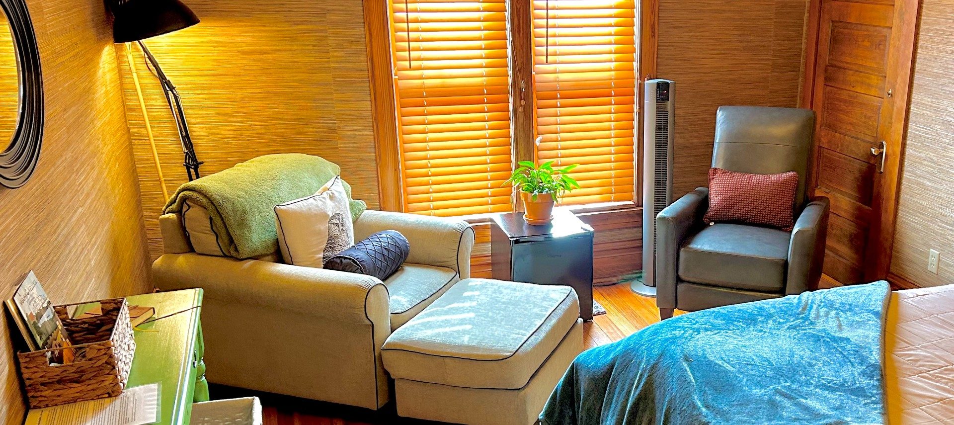 Sitting area off a bedroom with two upholstered sitting chairs, table with a plant by a large window with blinds