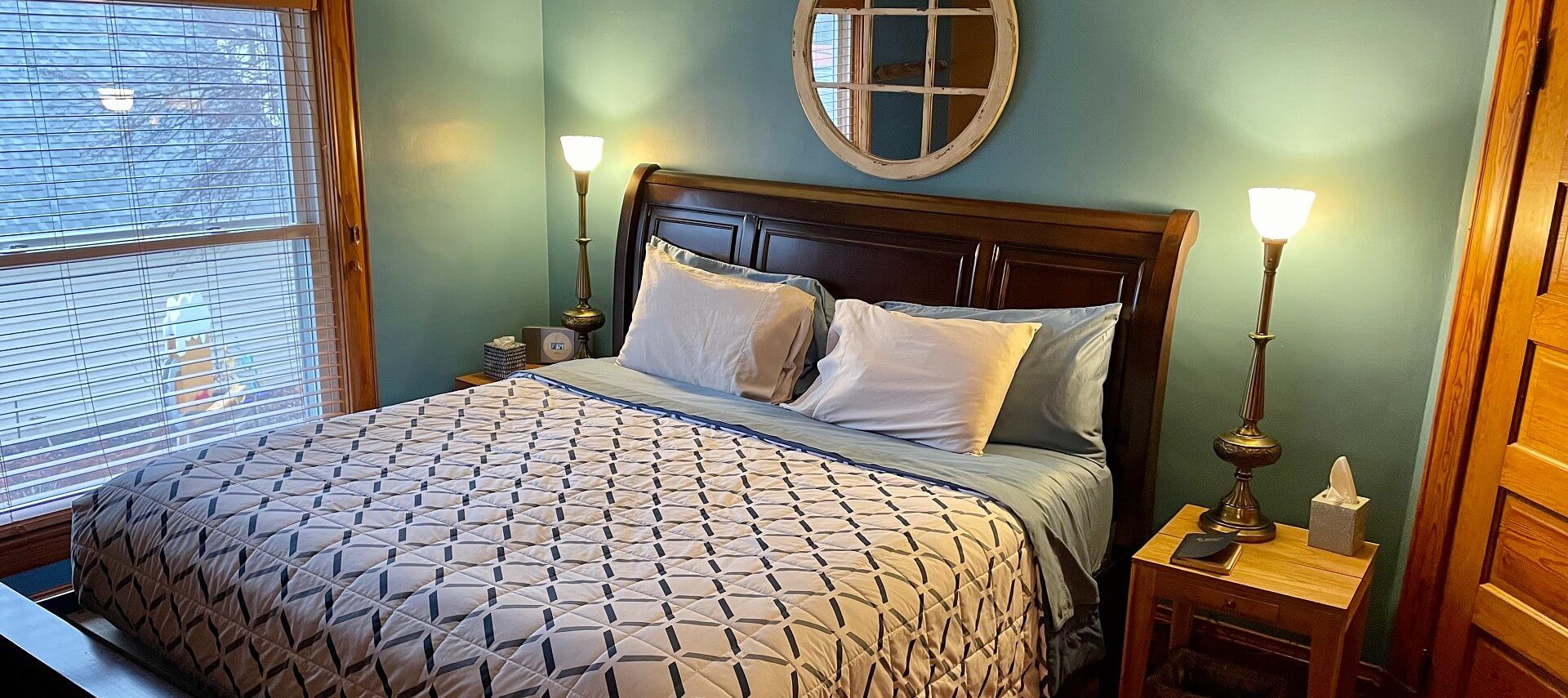 Green bedroom with king bed with dark wood sleigh headboard, side tables with tall lamps and large window with blinds