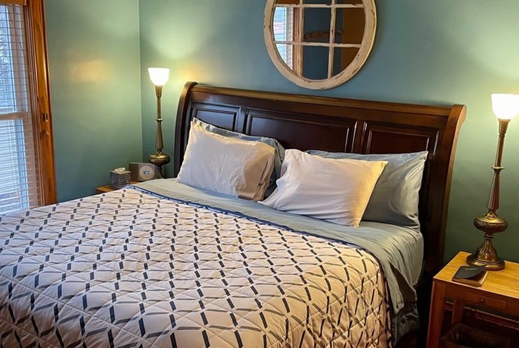 Green bedroom with king bed with dark wood sleigh headboard, side tables with tall lamps and large window with blinds