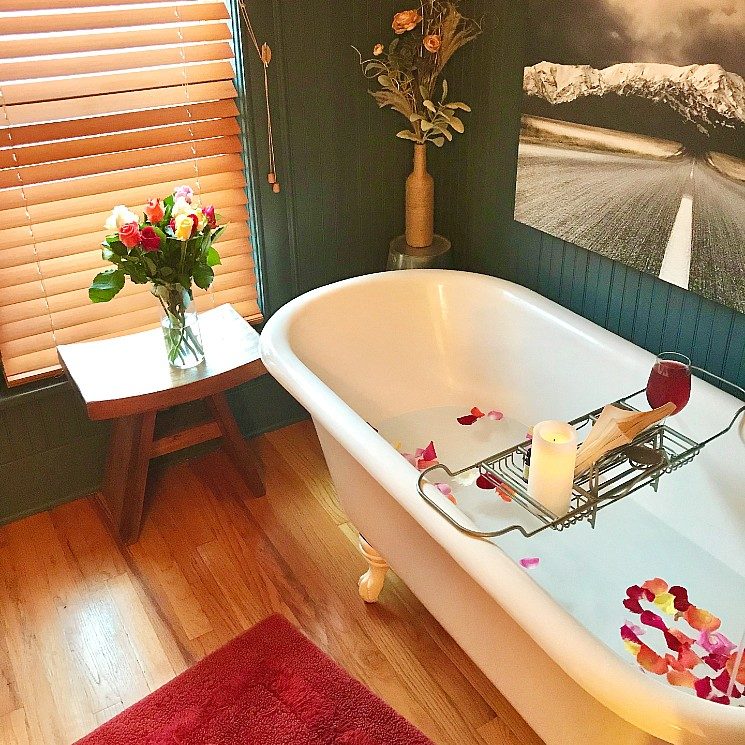 White clawfoot tub full of water and rose petals, stool with vase of flowers and artwork on the wall