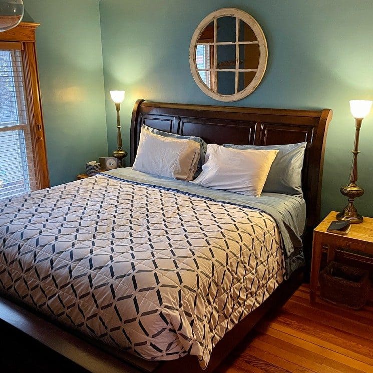 Green bedroom with king bed with dark wood sleigh headboard, side tables with tall lamps and large window with blinds