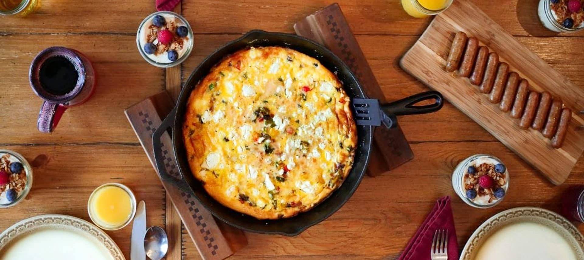 Wood dining able with cast iron pan with a breakfast dish, platter of sausage links and glasses of orange juice