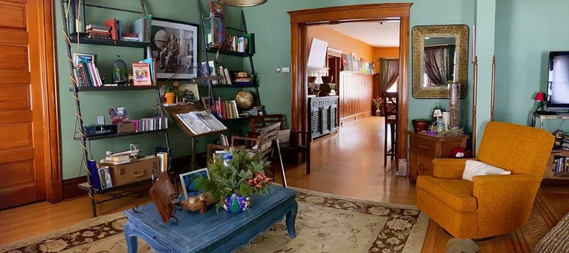 Living area of a home wth orange chair, blue coffee table, shelving unit on the wall and doorway into a separate room