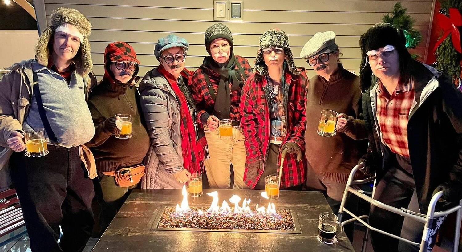 Seven girls dressed up as old men drinking beer around a firepit table